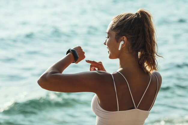 Una bella giovane donna sta guardando smartwatch e si prepara a fare jogging sulla spiaggia del mare in una giornata di sole estivo.