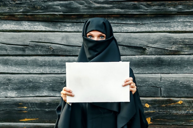 Una bella, giovane ragazza musulmana con un velo nero con la faccia chiusa contro un albero grigio. Nel foglio di carta handsa. copyspace.