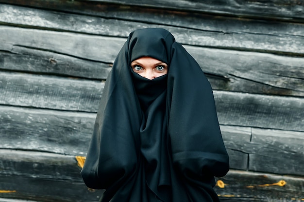 Una bella, giovane ragazza musulmana in un velo nero con la faccia chiusa su un albero grigio tiene le mani dietro la testa. copyspace.