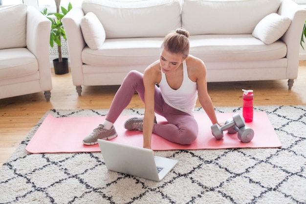 Una bella modella in abbigliamento da ginnastica seduta su un tappetino usando un portatile e preparandosi per un allenamento a casa