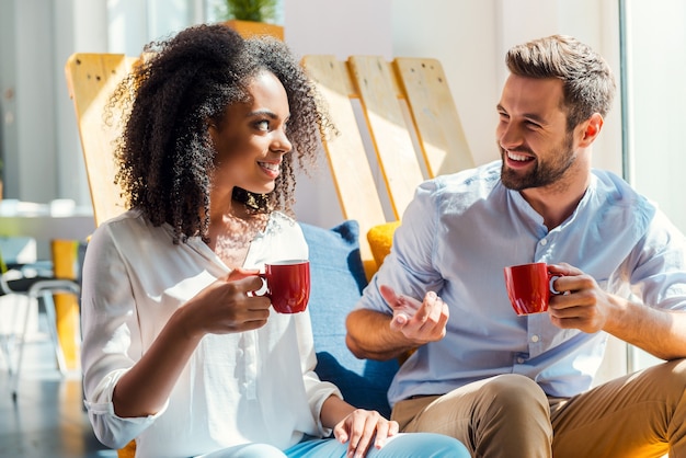 Una bella pausa caffè. Allegra giovane donna africana e giovane uomo che tiene tazze di caffè e discutono di qualcosa mentre sono seduti nell'area di riposo dell'ufficio