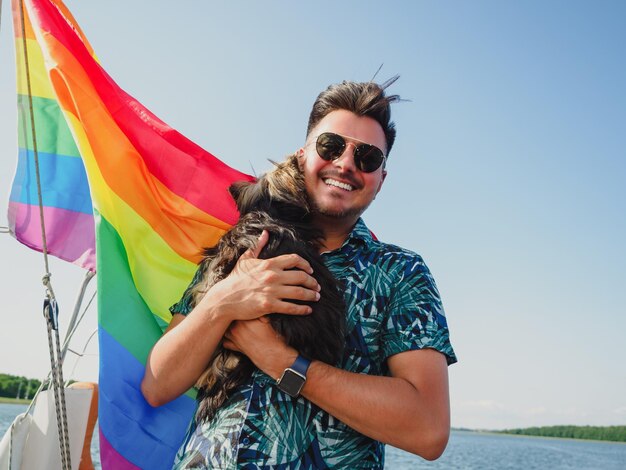 Una bella persona gay sta abbracciando il suo piccolo cane Yorkshire terrier su una bandiera arcobaleno sullo sfondo