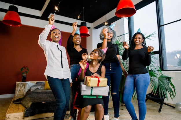 Una bella ragazza africana sorridente tiene regali nelle sue mani e festeggia il compleanno con gli amici. Le ragazze tengono in mano le luci del Bengala