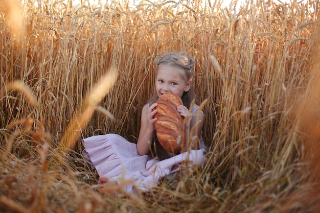 una bella ragazza bionda con un vestito di lino rosa è seduta in un campo di grano e mangia pane