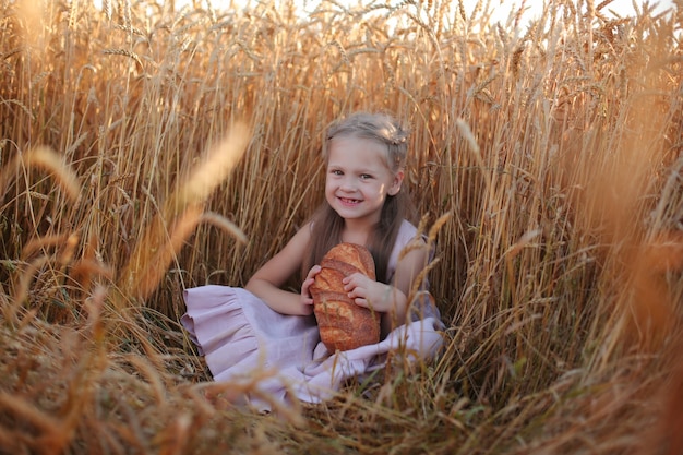 una bella ragazza bionda con un vestito di lino rosa è seduta in un campo di grano e mangia pane