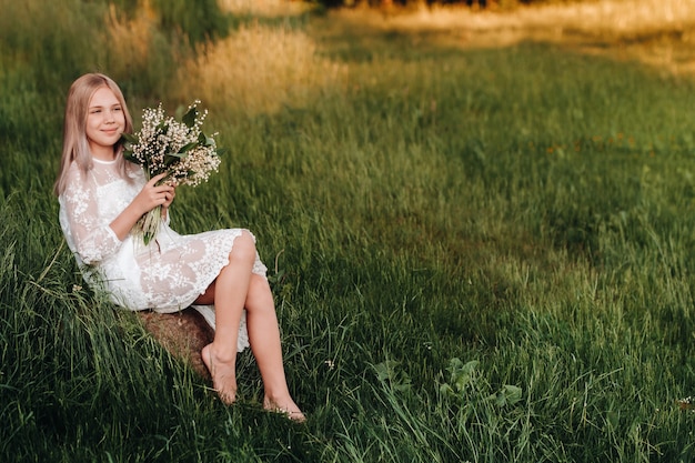 Una bella ragazza bionda di nove anni con i capelli lunghi in un lungo abito bianco, che tiene in mano un mazzo di fiori di mughetto, cammina nella natura nel parco. Estate, tramonto.