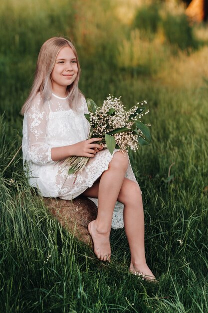 Una bella ragazza bionda di nove anni con i capelli lunghi in un lungo abito bianco, che tiene in mano un mazzo di fiori di mughetto, cammina nella natura nel parco. Estate, tramonto.