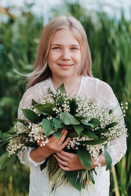 Una bella ragazza bionda di nove anni con i capelli lunghi in un lungo abito bianco, che tiene in mano un mazzo di fiori di mughetto, cammina nella natura nel parco. Estate, tramonto.