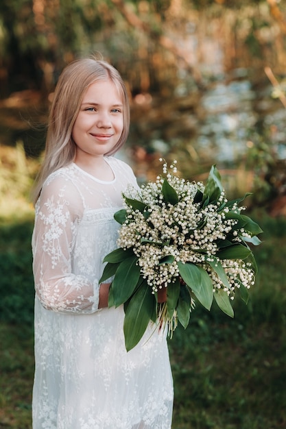 Una bella ragazza bionda di nove anni con i capelli lunghi in un lungo abito bianco, con in mano un mazzo di fiori di mughetto, passeggiando nella natura nel parco. Estate, tramonto.