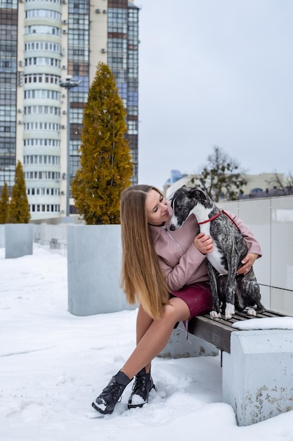 una bella ragazza bionda è seduta su una panchina e abbraccia un cane whippet