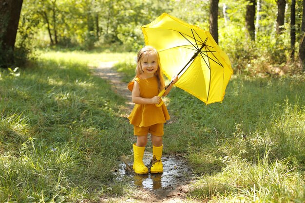 una bella ragazza bionda in un abito di lino con stivali gialli con un ombrello giallo nel parco