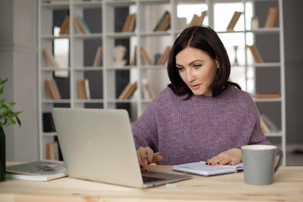una bella ragazza bruna con un maglione lilla è seduta a un tavolo con un laptop che prende appunti