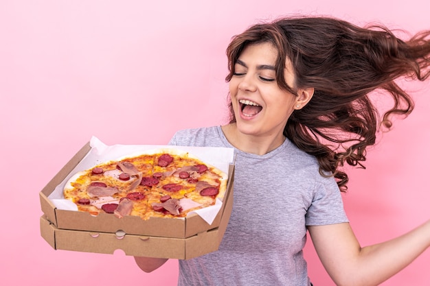 Una bella ragazza bruna si rallegra della pizza dalla scatola per la consegna su uno sfondo rosa, copia spazio.