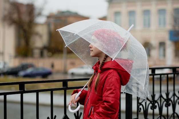 Una bella ragazza cammina sul ponte sotto la pioggia con un ombrello trasparente.