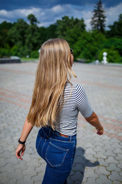 Una bella ragazza con i capelli lunghi biondi di aspetto europeo. Vestito con jeans e maglietta. Cammina in una giornata di sole