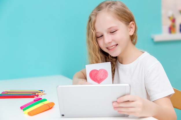 Una bella ragazza con lunghi capelli rossi e occhi azzurri in una maglietta bianca su sfondo color acquamarina disegna un cuore rosso con una matita per San Valentino e comunica online su un tablet showi