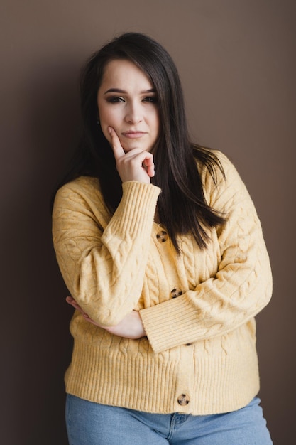 Una bella ragazza con lunghi capelli scuri in un maglione giallo si erge pensierosa su uno sfondo marrone