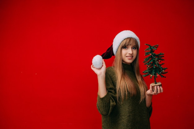 Una bella ragazza con un cappello da Babbo Natale tiene in mano un piccolo albero di Natale su uno sfondo rosso