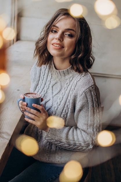 Una bella ragazza con un maglione beve tè o caffè sullo sfondo di una finestra e delle luci