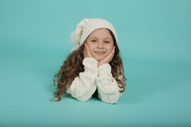 una bella ragazza con un maglione bianco e un cappello invernale bianco giace su uno sfondo blu