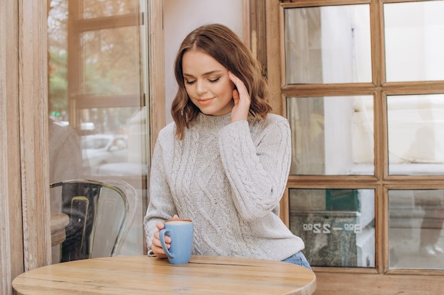 Una bella ragazza con un maglione grigio beve caffè mattutino o cacao in una caffetteria Riposo autunnale