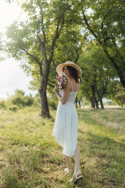 Una bella ragazza con un prendisole bianco e un cappello sta camminando lungo una strada forestale con fiori in mano Il concetto di vacanza estiva