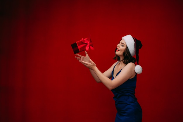 Una bella ragazza con un vestito blu e un cappello da Babbo Natale prende un regalo di Natale su uno sfondo rosso