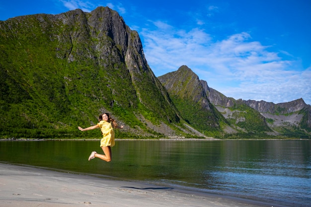 una bella ragazza con un vestito giallo salta di gioia su una spiaggia circondata da possenti montagne, senja