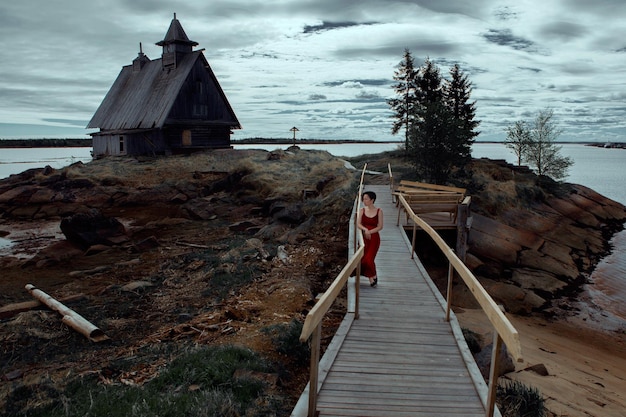 una bella ragazza con un vestito rosso sta da sola sulla riva su un ponte vicino a una casa di legno abbandonata