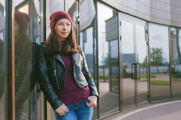 Una bella ragazza con una giacca di pelle e un cappello si erge sullo sfondo di un edificio di vetro.