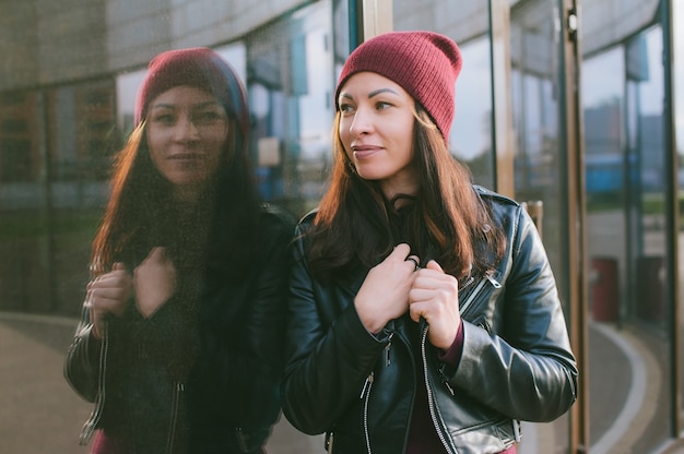 Una bella ragazza con una giacca di pelle e un cappello si erge sullo sfondo di un edificio di vetro.