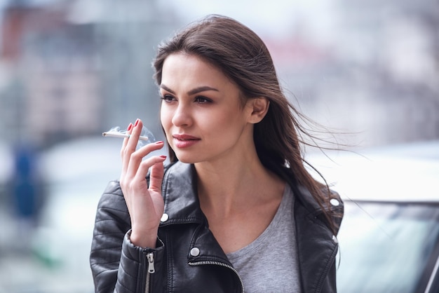 Una bella ragazza con una giacca di pelle sta fumando e guarda lontano mentre si appoggia sulla sua macchina