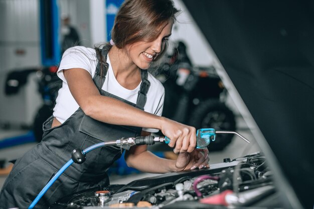 Una bella ragazza con una tuta nera e una maglietta bianca sta sorridendo, controllando il livello dell'olio in un'auto nera nel garage.