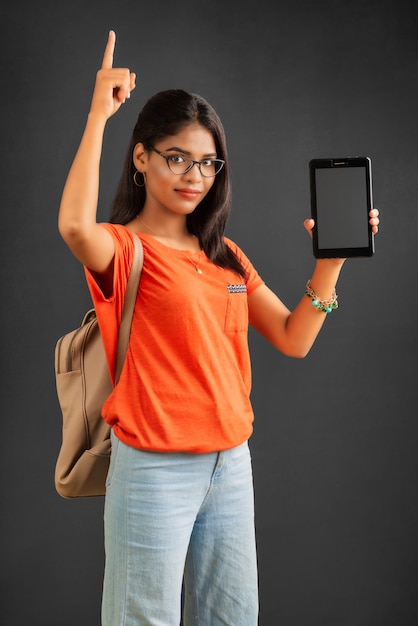 Una bella ragazza con uno zaino mostra uno schermo vuoto di uno smartphone o un telefono cellulare o tablet in posa su uno sfondo grigio