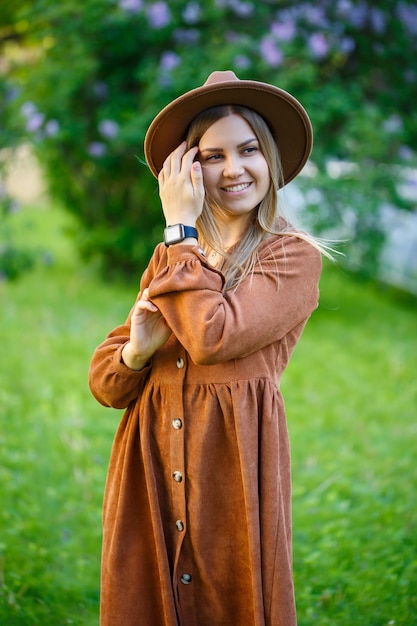 Una bella ragazza dai capelli biondi tiene in mano un cappello e sta con gli occhi chiusi vicino a un cespuglio di lillà. Giovane donna in un giardino con alberi in fiore