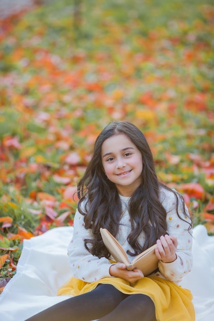 Una bella ragazza di 10 anni dai capelli scuri con un maglione lavorato a maglia legge un libro seduto in una radura in un parco autunnale Picnic nella natura Il bambino felice sta imparando