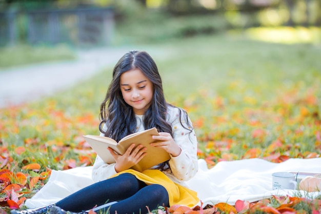 Una bella ragazza di 10 anni dai capelli scuri con un maglione lavorato a maglia legge un libro seduto in una radura in un parco autunnale Picnic nella natura Il bambino felice sta imparando