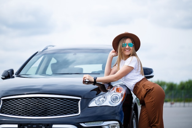 Una bella ragazza di aspetto europeo con gli occhiali e un cappello marrone si trova vicino a un'auto nera. Servizio fotografico vicino alla macchina.