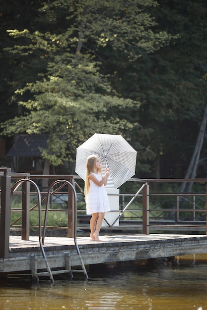 Una bella ragazza di sei anni con un abito corto bianco si erge sulla muratura vicino al fiume e tiene in mano un ombrello bianco