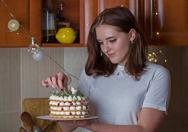 Una bella ragazza felice con una maglietta bianca sta a casa tiene una torta di Capodanno festiva su un piatto