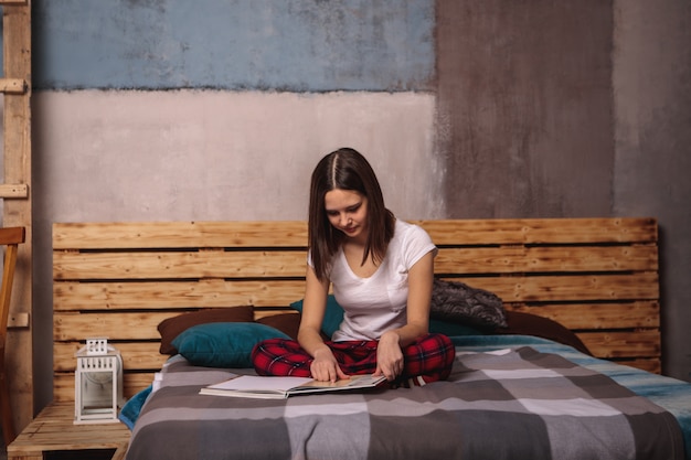 Una bella ragazza in abiti domestici è seduta sul letto e legge un libro, guardando una rivista. Hobby, tempo libero, tempo libero. Lo sguardo è diretto verso il basso nel libro.