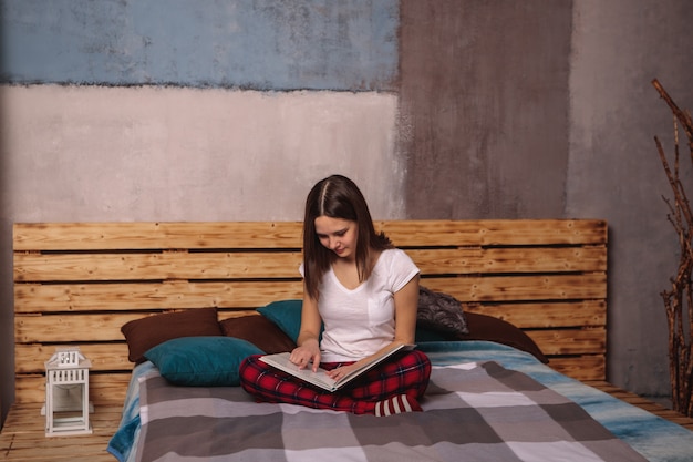 Una bella ragazza in abiti domestici è seduta sul letto e legge un libro, guardando una rivista. Hobby, tempo libero, tempo libero. Lo sguardo è diretto verso il basso nel libro.