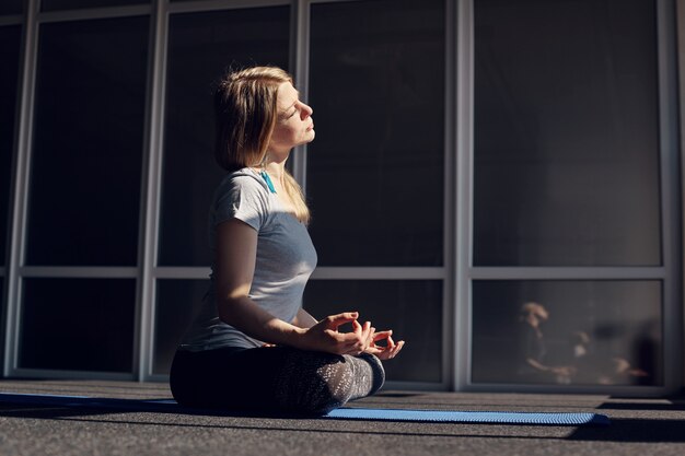 Una bella ragazza in abiti sportivi medita durante le lezioni di yoga. La bionda è seduta nella posizione del loto, rilassata, esponendo il viso al sole. Il concetto di uno stile di vita sano. Vista laterale