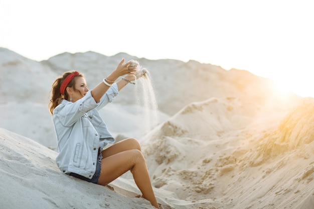 Una bella ragazza in jeans, pantaloncini corti, un cerchio rosso e un reggiseno bianco è seduta sulla sabbia