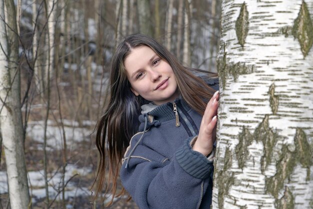 Una bella ragazza in primavera in natura guarda fuori da dietro un albero di betulla.