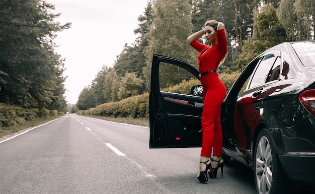 Una bella ragazza in tuta rossa si trova accanto a un'auto nera su una strada deserta nella foresta