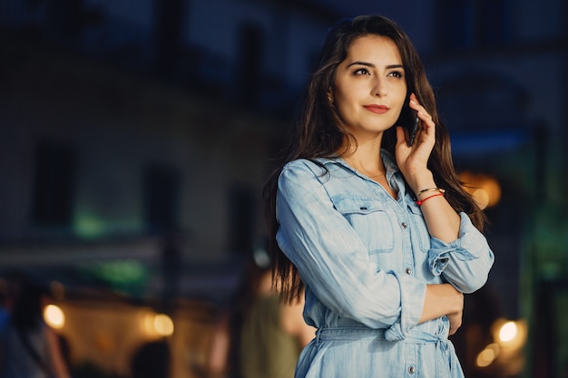 Una bella ragazza in un abito blu in piedi in una città di notte con il telefono