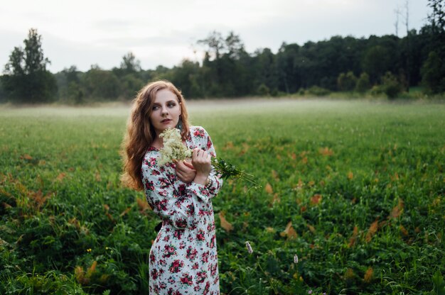 Una bella ragazza in un abito colorato si trova la sera su un prato su uno sfondo di nebbia.