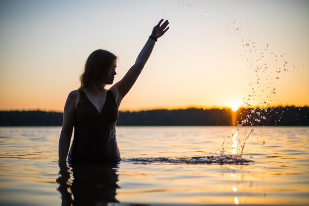 Una bella ragazza in un lungo costume da bagno nero nuota sul lago nei raggi del tramonto o dell'alba