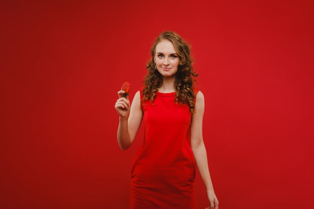 Una bella ragazza in un vestito rosso su sfondo rosso tiene una fragola tra le mani e sorride.